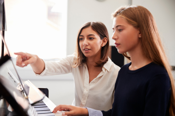 two young women sitting at a piano. The piano teacher points to the music while the piano student plays