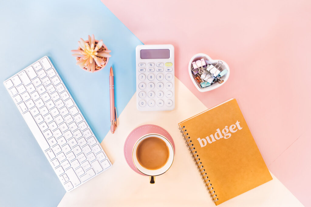 a keyboard, calculator, coffee, and budget notebook on a blue and pink background to calculate the hidden costs of being a music teacher